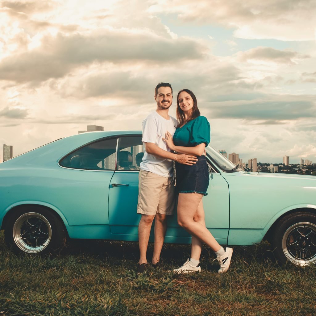 Happy couple standing beside a vintage blue car with city skyline in the background at sunset.