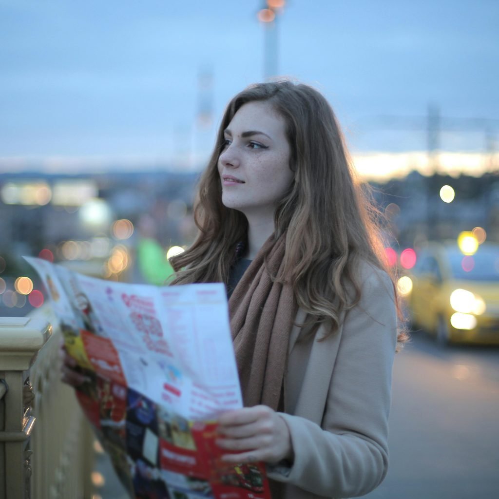 Female traveler in warm clothes standing on street with map and looking away at dusk